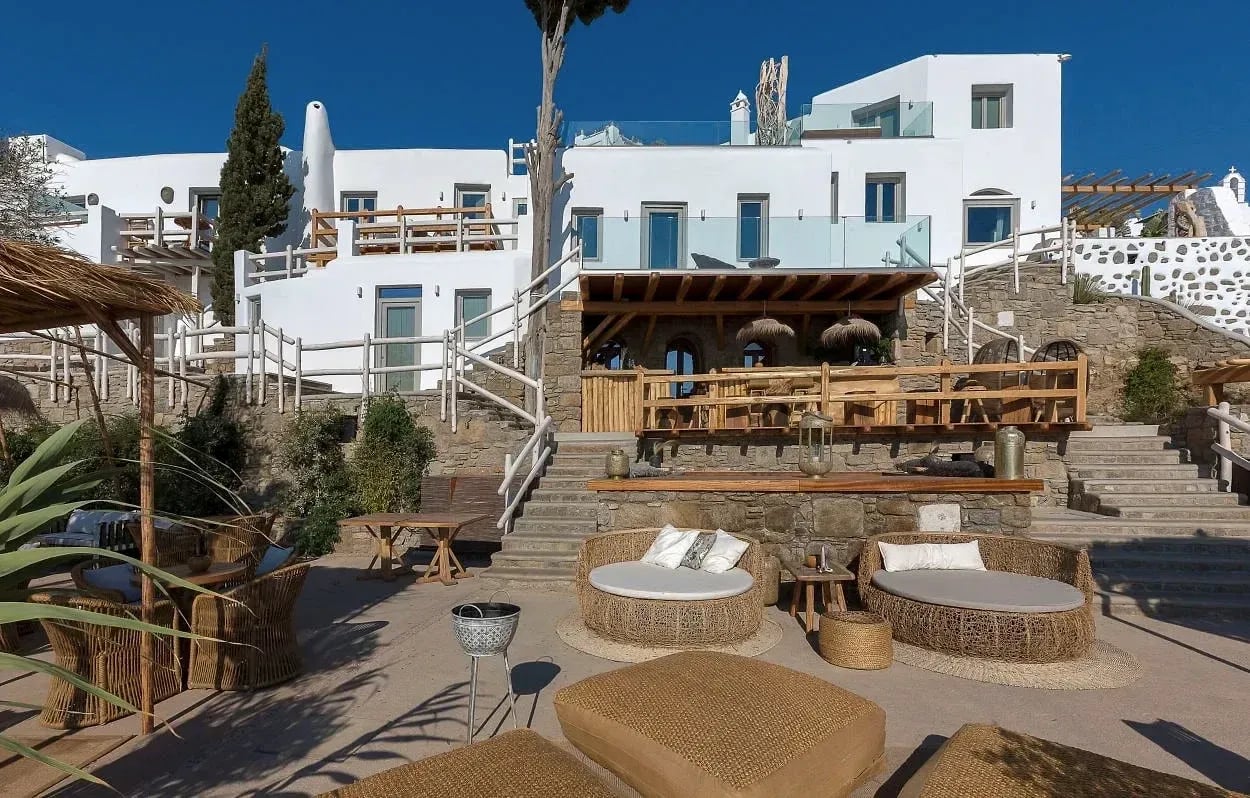 The Garden of Mykonos bar below the suites, with timber counter, straw parasols and woven floor seating